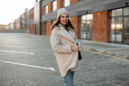 Pretty funny young woman in a fashionable knitted hat in a stylish coat of milk color from eco-fur stands and smiling near a brick vintage building. Cheerful girl model resting outdoors. Spring look.の写真素材