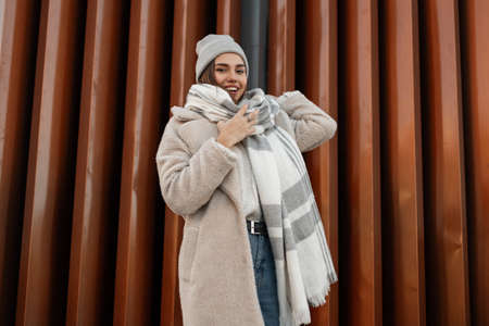 Urban cute funny young woman in fashionable knitted clothes posing near a metal wall in the city. Pretty happy girl fashion model with a positive smile enjoys a rest outdoors in a warm spring day.の写真素材