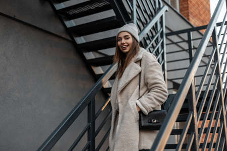 Happy pretty young woman with cute smile in stylish warm seasonal outerwear stands and smiles on a vintage metal staircase near a building on the street. Joyful girl in trendy clothes in the city.の写真素材