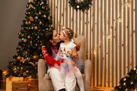 Pretty happy mother with cute daughter sits and kisses on the cheek in the New Year's room with festive decorations. Young woman with a beautiful little girl is resting on a chair in the living room.の写真素材
