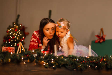 Young mother in a knitted trendy red sweater with a cute small daughter in a funny unicorn costume with a full skirt blow out candles in a room with a Christmas tree. family weekend. Happy New Year.の写真素材