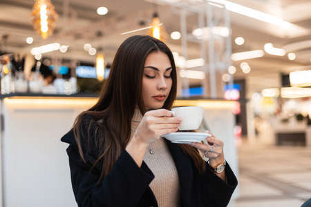 Stylish young beautiful brunette woman with long hair in black fashionable coat with cup of coffee is resting in cafe in mall. Cute attractive modern girl in trendy clothes is enjoying hot drink indoorsの写真素材
