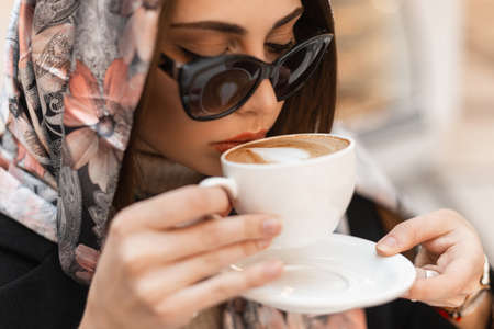 Portrait trendy young luxurious woman in fashionable silk shawl on head in stylish dark sunglasses with a vintage white cup with coffee. Attractive fashion model girl drinks hot cappuccino. close-up.の写真素材