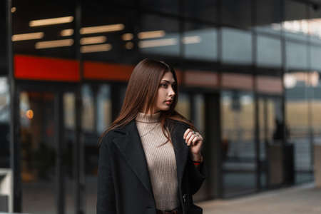 Urban pretty stylish young woman model in a fashionable black coat in a beige shirt stands near a vintage glass building in the city. European modern girl in elegant clothes outdoors. youth style.の写真素材