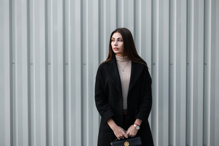 Fashionable young woman with brown hair in black fashionable coat with stylish leather black handbag stands near silver metal vintage wall. Modern luxurious girl fashion model outdoors. trendy lady.の写真素材