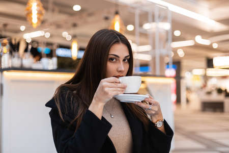 Fashionable young woman with long hair in black stylish coat sits and drinks hot tasty tea in a cafe...