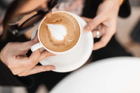 Top view a cup of hot cappuccino with foam in a white vintage cup in female hands. Fashionable young woman in stylish sunglasses enjoys caffeine in the morning at a cafe. close-up.の写真素材
