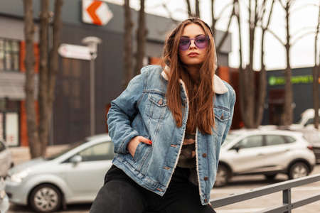 Fashion model young beautiful woman in fashionable oversized blue denim jacket in hood with purple glamorous glasses posing near on the street in the city. Hipster urban girl outdoors. american styleの写真素材