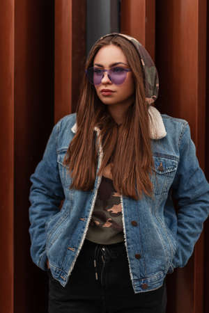 Glamorous beautiful young hipster woman in trendy purple glasses in oversized blue jeans jacket with long hair is rests near metal wall in the city. Modern urban girl model in stylish clothes outdoorsの写真素材