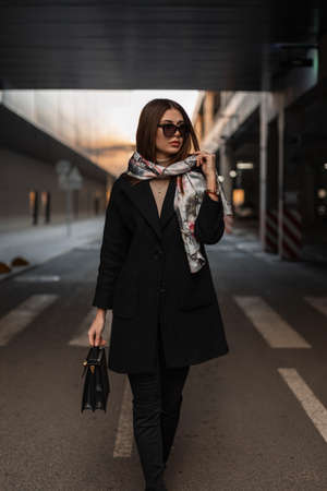 Pretty business young woman model with elegant scarf on head in fashionable sunglasses in a trendy coat with leather black handbag stands on road in the city. Sexy girl posing outdoors. modern lady.の写真素材