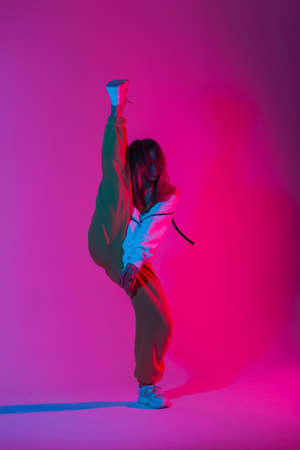 Fashion model young modern woman in stylish clothes in trendy gym shoes dancing indoors with bright multi-colored color. Sporty girl dancer posing in a room with bright neon glamor pink-blue light.の写真素材