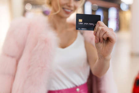 Close-up young female face with charming smile with teeth. Attractive happy woman in pink fur coat in fashion white t-shirt smiles cute holding credit card in hands for shopping.の写真素材