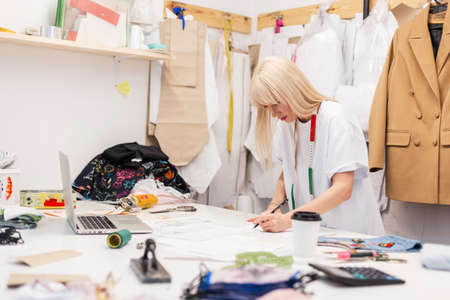 Young fashion designer woman draws a sketch on desktop with a laptop, coffee, threads, centimeter,...