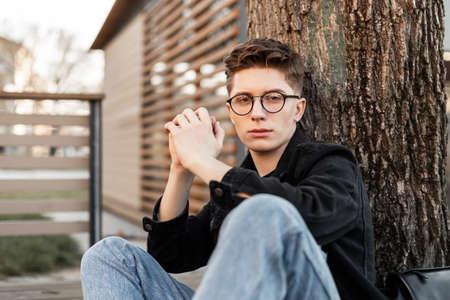 Fresh portrait serious young hipster man in fashionable glasses in denim jacket in blue jeans with...
