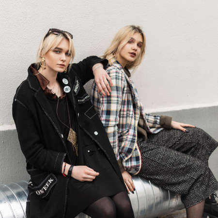 Two attractive young blonde girlfriends in oversized stylish clothes in retro style sit on silver metal pipe near wall on street. Sexy girls in trendy youth outfit. Gorgeous fashion models outdoors.の写真素材