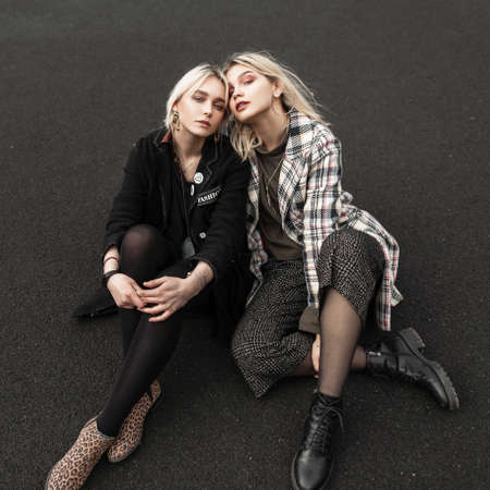 Fashion lovely girlfriends in fashionable jackets in cool stylish shoes resting on asphalt on street in city. Gorgeous beautiful teenage girls in trendy clothes sitting outdoors. New spring youth wearの写真素材
