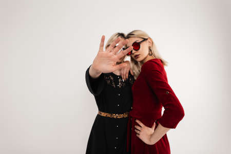 Pretty modern fashionable teenage girls in fashion clothes in stylish colored glasses hugging and lady in black dress closes camera with hand. Trendy sisters in elegant attire posing in studio. Retroの写真素材