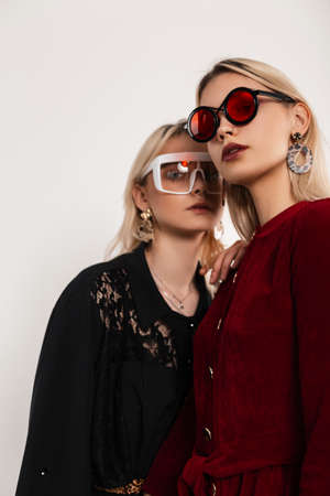 Fresh portrait young beautiful lesbians in fashionable red-black clothes in stylish cool glasses stand next to each other on gray background indoors. Trendy girls in elegant dresses posing in studio.の写真素材