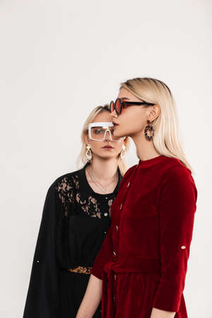 Fashion portrait young attractive blondes sisters women in red-black fashionable dresses in stylish glasses near gray vintage wall in studio. Sexy stylish girls posing indoors. Lovely elegant ladies.の写真素材