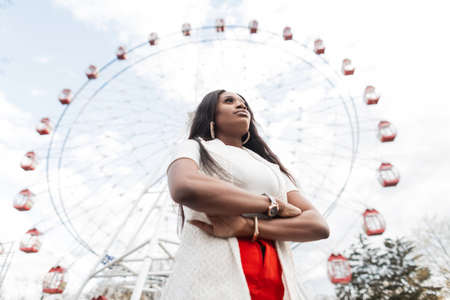 Elegant modern sexy black young woman in elegant clothes stands against the sky and ferris wheel. Beautiful fashionable african girl travels on city near amusement park. Stylish wear for women.の写真素材