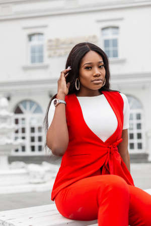 Modern fashionable young black woman in beautiful stylish red suit is resting on wooden bench in...
