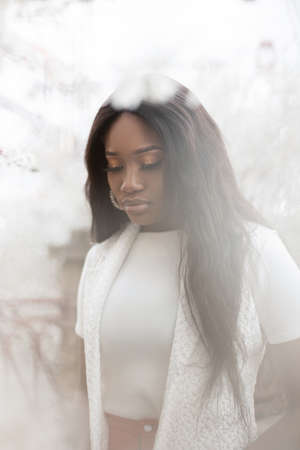 Cute beautiful tender young black woman in a white dress on a background of white flowers outdoors....