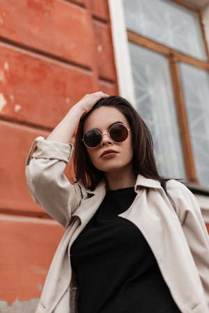 Portrait attractive young woman fashion model in fashionable sunglasses with sexy lips in spring clothes outdoors near vintage building. Beautiful stylish girl walks on street and straightens hair.の写真素材