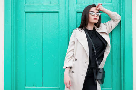 Sexy young woman in stylish retro sunglasses in black trendy t-shirt in fashionable spring trench coat is rest near vintage bright wooden turquoise door on street. Attractive girl model in the city.の写真素材