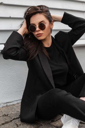 Lovely fashion pretty young woman in sunglasses in fashionable black casual wear straightens hair near vintage wooden building on street in city. American trendy girl model is resting on stone tile.の写真素材
