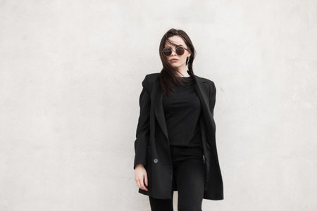 Trendy model gorgeous young woman in cool mirror sunglasses in fashionable black casual jacket in stylish t-shirt near modern white wall in city. Urban sexy girl in fashion youth outfit in street.の写真素材