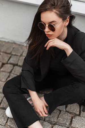 Young beautiful woman in trendy sunglasses in cool black youth clothes is resting near vintage building in city. Fashionable girl enjoys a rest on stone tile on street. Spring fashion wear for women.の写真素材
