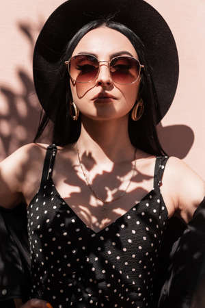 Stylish sexy young woman in hat in jacket in dress in sunglasses enjoys sun on street. Fashion portrait vogue girl in black elegant summer beautiful clothes in bright sunlight near vintage pink wall.の写真素材