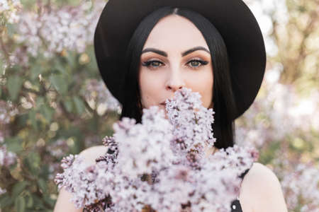 Pretty young woman with beautiful eyes in elegant stylish hat holds near face bouquet fresh lilac flowers on background flowering trees. Cute girl walks on sunny day in park and enjoys spring and sun.の写真素材