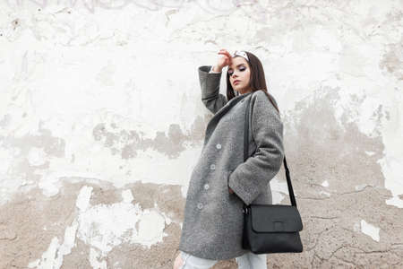Fashion model cute woman in gray elegant coat in glamorous bandana with stylish black bag relaxes near vintage white wall in city.Gorgeous girl in casual outerwear enjoys spring day on street outdoorsの写真素材