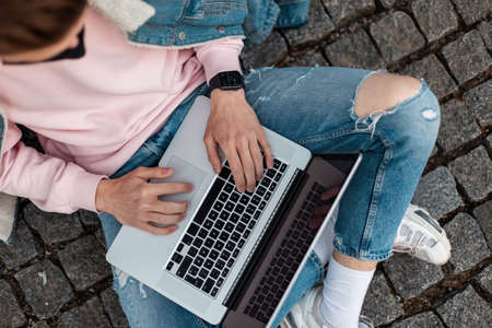 Man in medical black mask in jeans fashionable casual blue wear with laptop sits outdoors. Blogger professional works online on new creative project. Top view freelancer conceptの写真素材