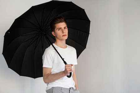 Fashion model handsome young man in youth fashionable T-shirt stands with vintage umbrella near wall in room. Sexy modern nice guy in summer wear posing with black umbrella on white background indoorsの写真素材