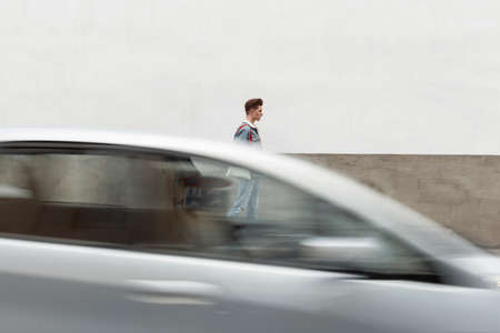 Young handsome man in stylish blue youth denim clothes is walking on street near road. Fashionable guy with casual jeans walks around the city near motion carの写真素材
