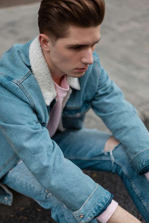 Fashionable young good-looking man in stylish blue jeans clothes resting on tile on street. Handsome guy with trendy hairstyle in fashion youth denim outfit sits in city near road. Casual wear for menの写真素材