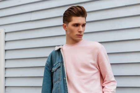 Handsome cute young man model in trendy pink sweatshirt in fashion denim blue youth jacket posing near white wall outdoors. Urban guy in stylish jeans casual wear from new collection poses on street.の写真素材