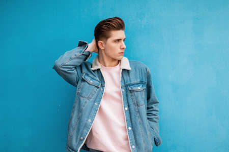 European young man in fashionable denim jacket in pink sweatshirt straightens stylish hairstyle near...