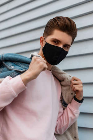 Portrait young man in denim blue stylish jacket in pink sweatshirt in black protective mask on street. Fashionable guy is walking in mask on city.の写真素材