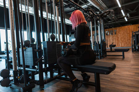 Beautiful fashionable athletic girl with pink hair in fashionable black sportswear sits near the simulator and prepares to do exercises in the gymの写真素材