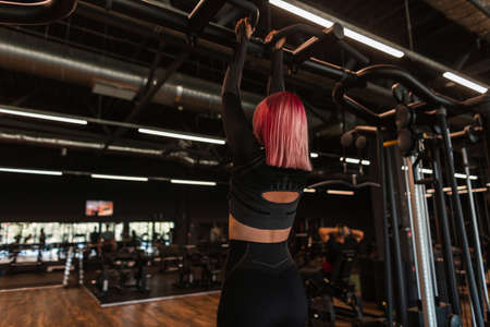 Stylish beautiful young girl athlete with pink hair in black sportswear training and exercising in the gymの写真素材