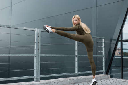 Beautiful sports fitness model woman in green workout clothes with white sneakers doing stretching exercise and warm up on a metal fence near a modern building. healthy lifestyleの写真素材