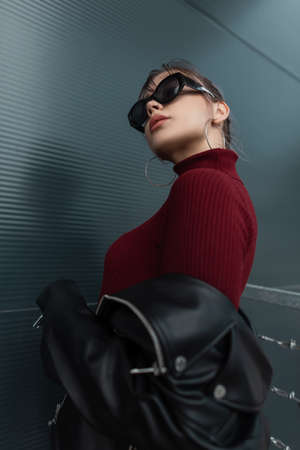 Fashionable portrait of a beautiful young woman in a burgundy sweater with a black leather jacket in sunglasses stands near a metal wall outdoorsの写真素材