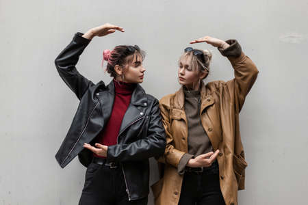 fashionable young two girlfriends girls with in cool leather jacket posing near a gray wall on the...