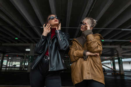 two beautiful young girls in youth style with leather jacket and black jeans with sunglasses posing in a parking lot on the streetの写真素材