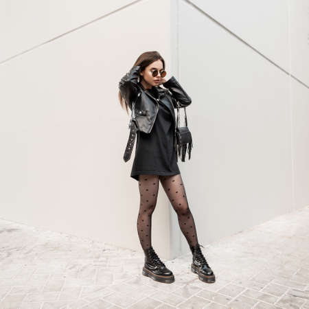 Pretty young cool hipster woman in black fashion rock outfit with leather jacket, sweatshirt, tights and shoes with a stylish black bag walks near a gray wall in the city. Urban female clothesの写真素材