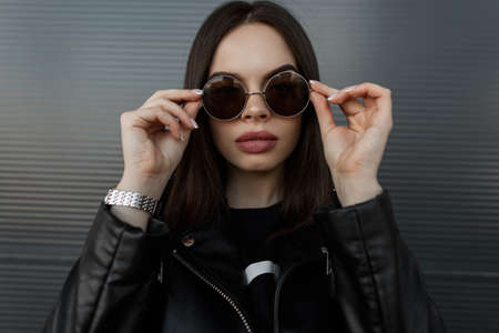 Fashion portrait of a beautiful hipster woman model with big natural lips in a stylish black leather jacket puts up cool round sunglasses near the metal wall on the streetの写真素材