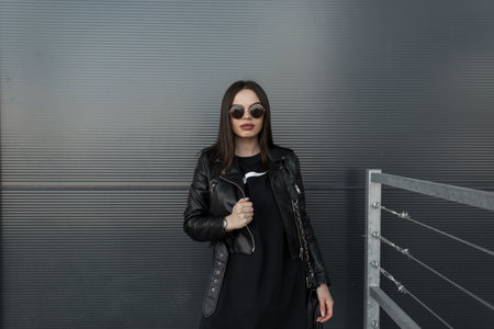 Stylish pretty hipster woman in fashionable round sunglasses and leather black jacket with sweatshirt stands near metal wall in cityの写真素材